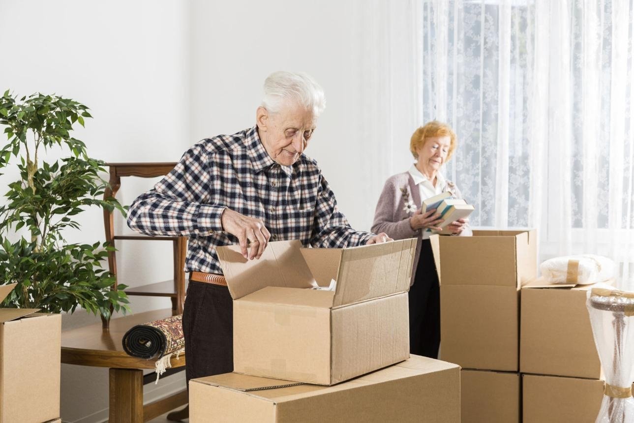 An elderly couple packing boxes in a bright living room, symbolizing downsizing to a simpler home.